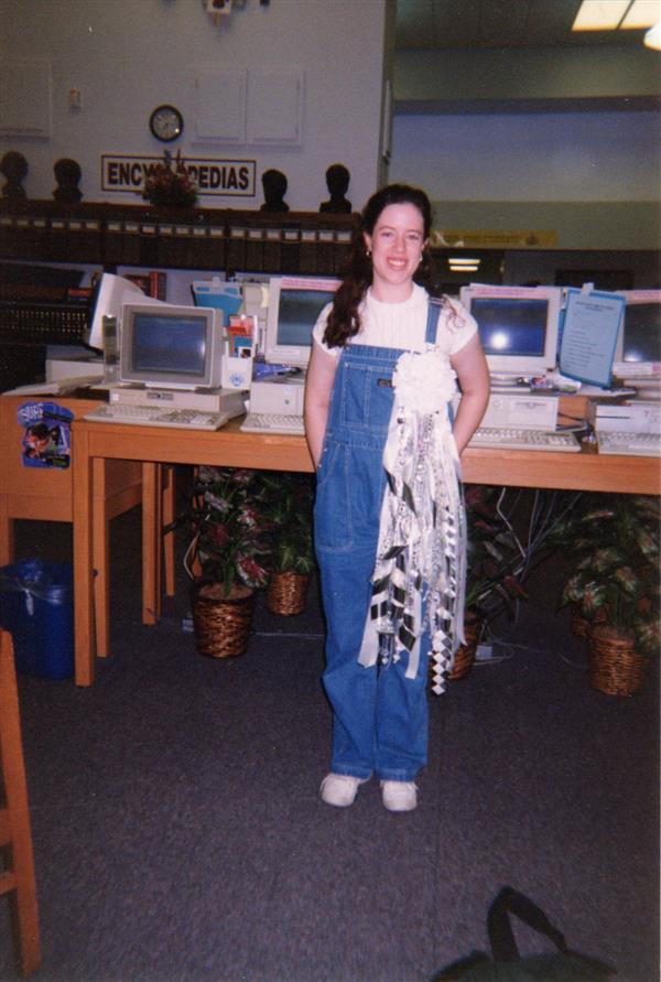 A teenage girl with long, dark hair is standing in front of computers in the PSHS library. She is wearing a white t-shirt under denim overalls and a Homecoming mum. The teenager is Mrs. Hoffmann when she was a senior here.